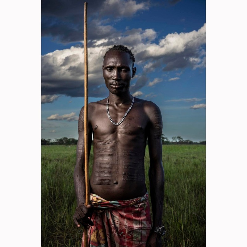 Người đàn ông cầm gậy đứng trên đồng cỏ Nam Sudan, với hoa văn nổi trên ngực và bụng